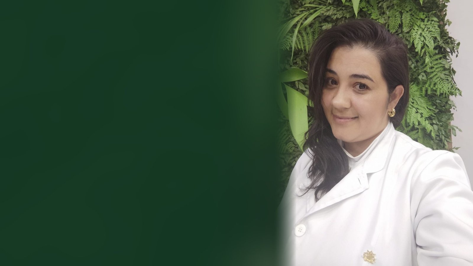 Tatiane Cubos, podóloga, de jaleco branco sorrindo em frente a um jardim vertical de folhagens
